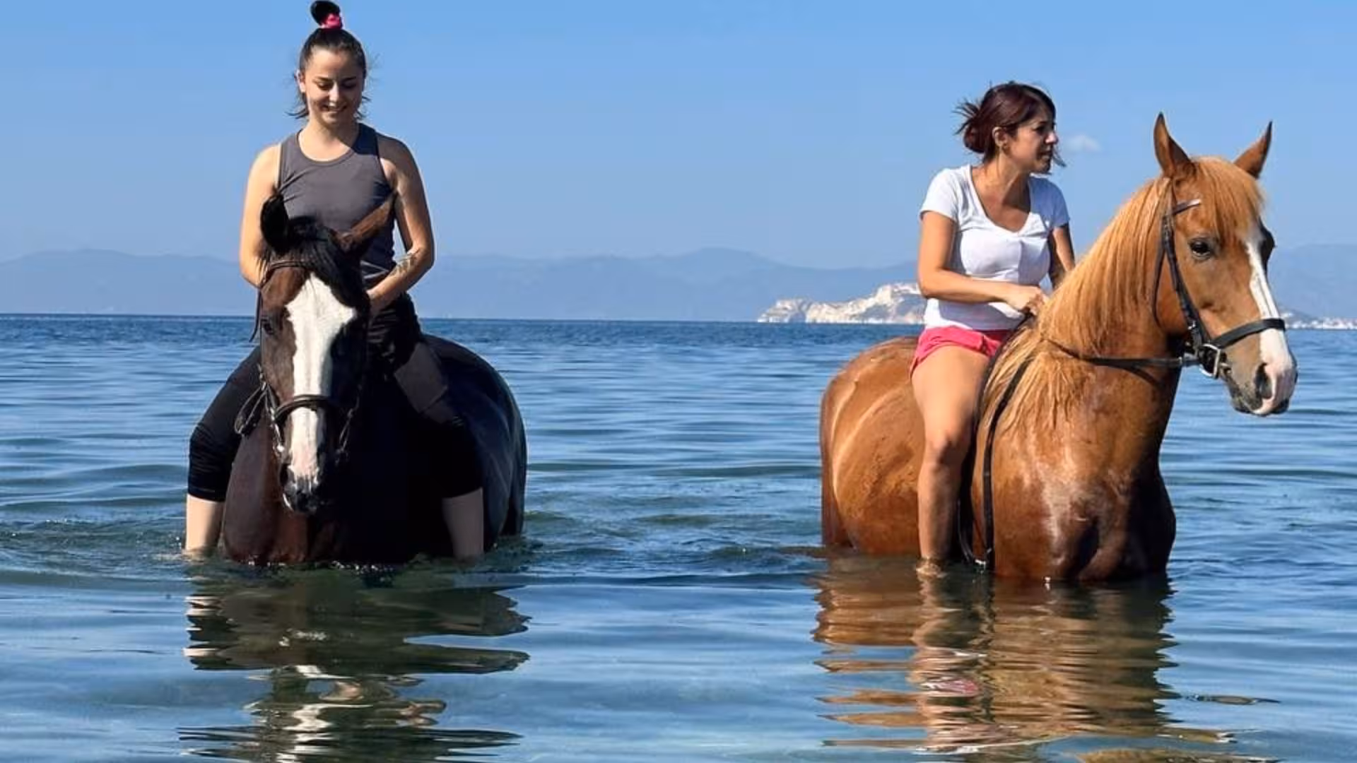 Two women enjoying horseback riding in the serene waters of Cagliari with distant mountains.
