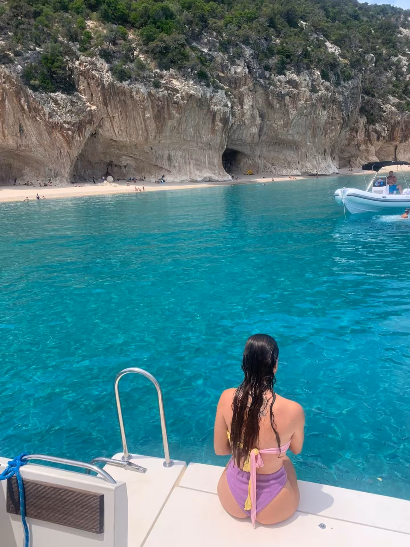 Woman in purple bikini sits on boat admiring Cala Gonone's crystal-clear waters and rocky coastline in the Gulf of Orosei.