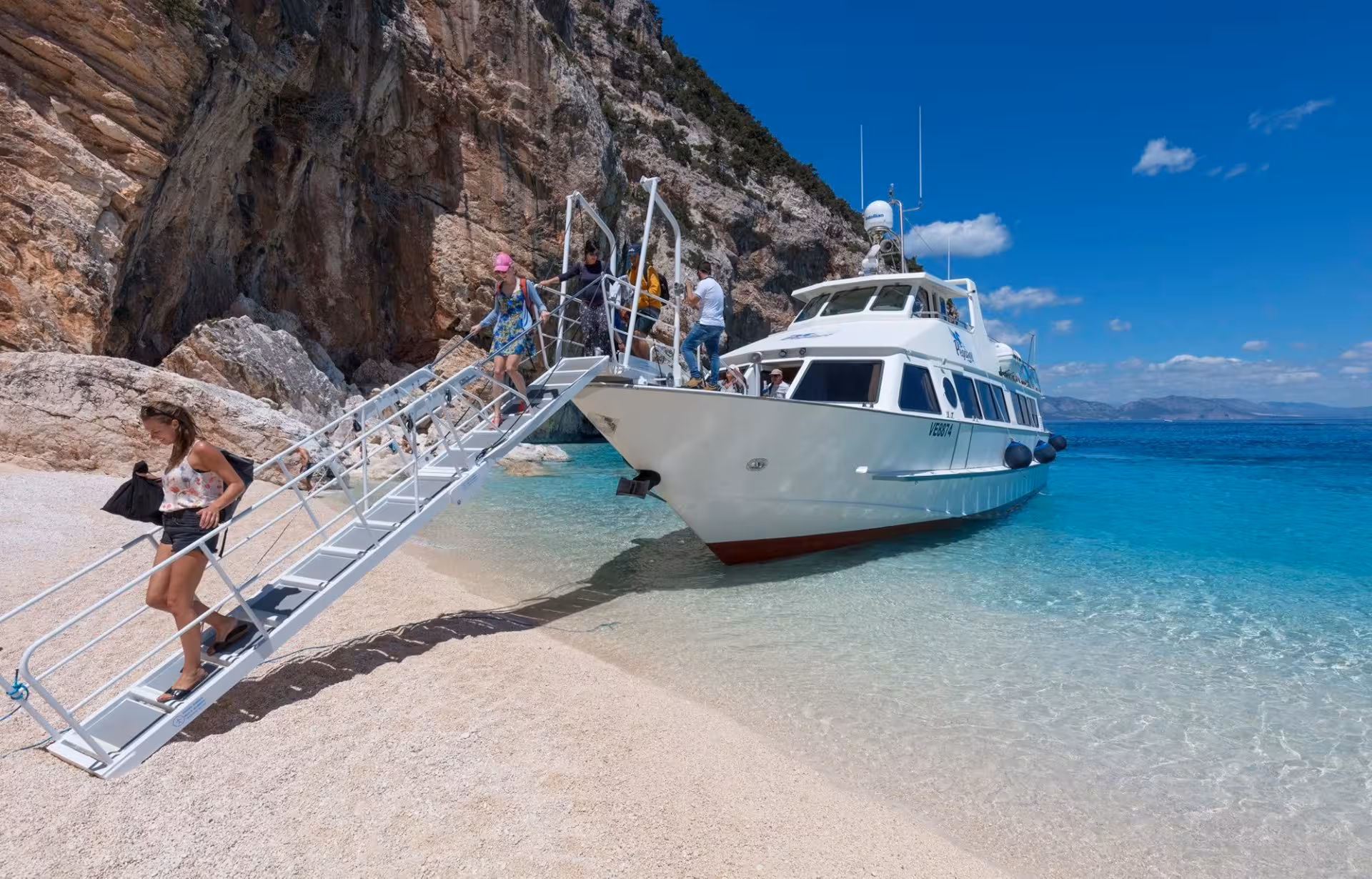 Tourists disembark from motorboat onto pristine beach during Cala Gonone tour to Bue Marino Caves.