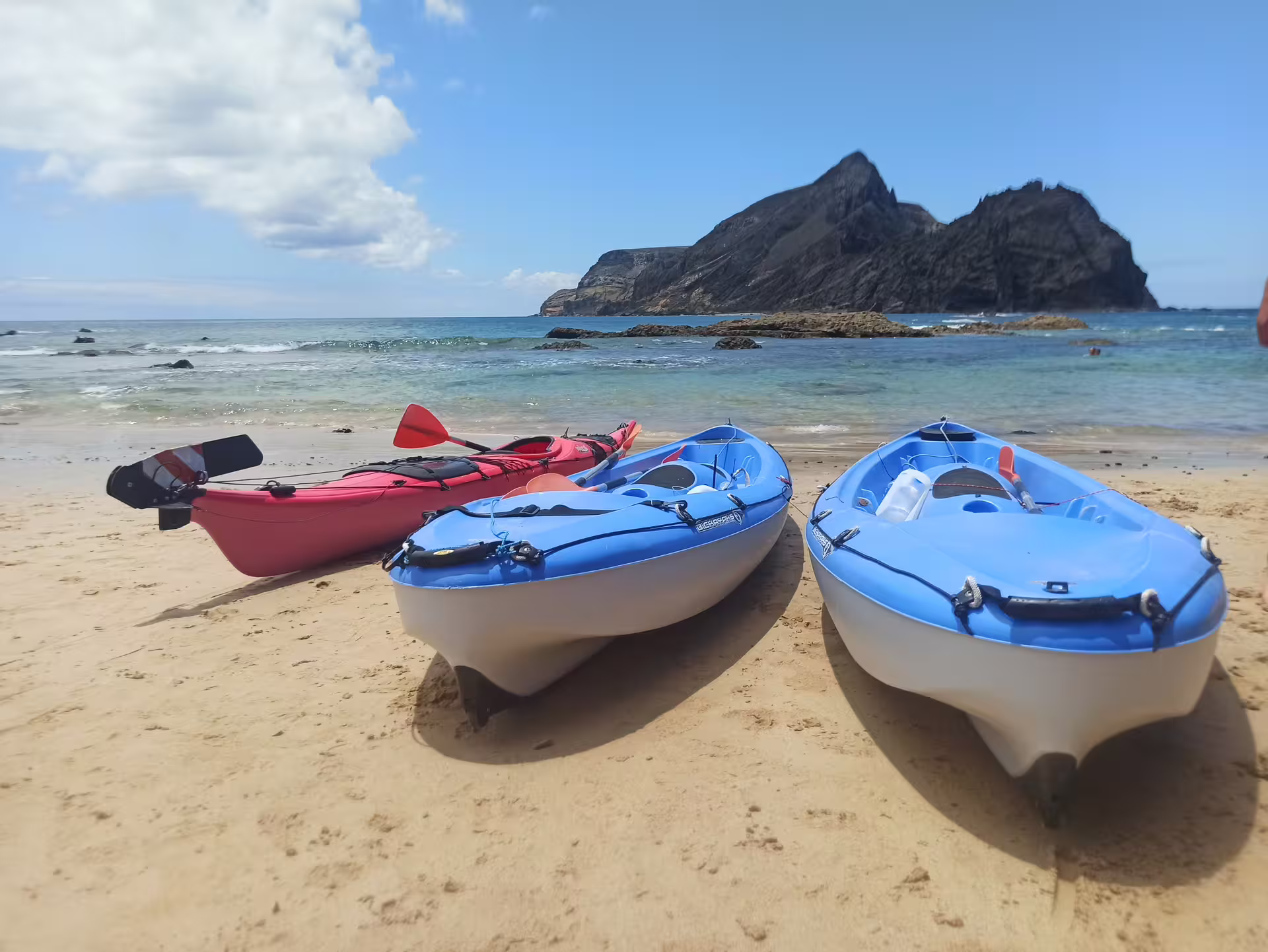 Three colorful kayaks on a sandy beach in Calheta with clear blue waters and rocky cliffs, perfect for an adventurous tour.