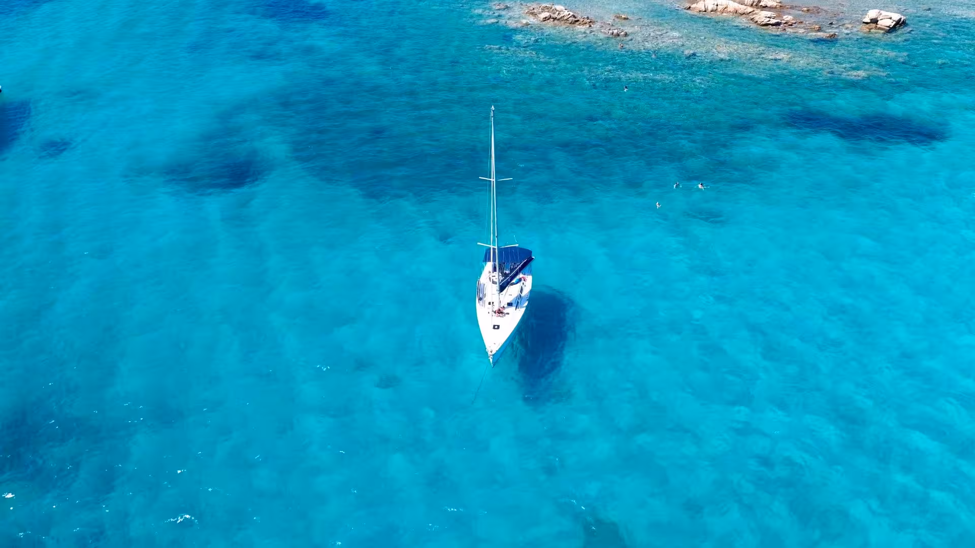 Aerial view of a sailboat with people enjoying the clear blue waters of La Maddalena Archipelago on a sunny day.