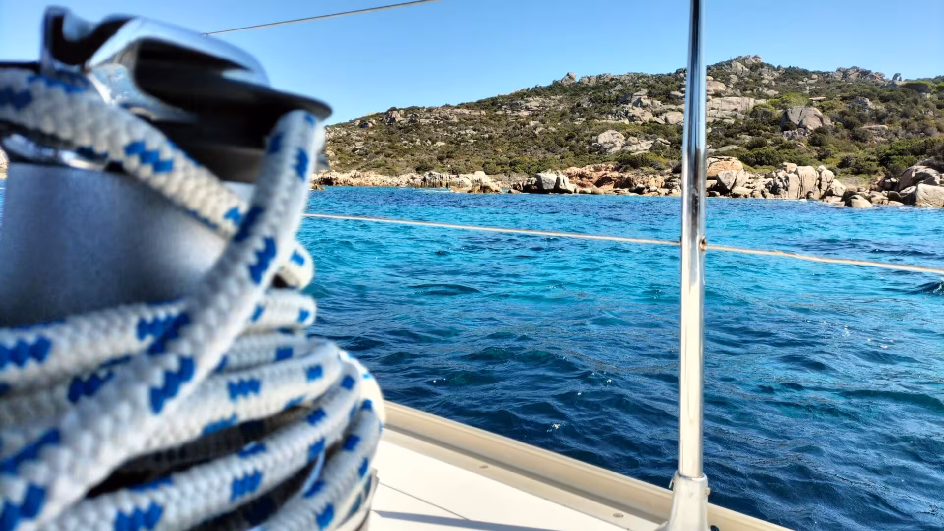 Close-up of sailing ropes with the turquoise waters and rocky coastline of La Maddalena Archipelago in the background.