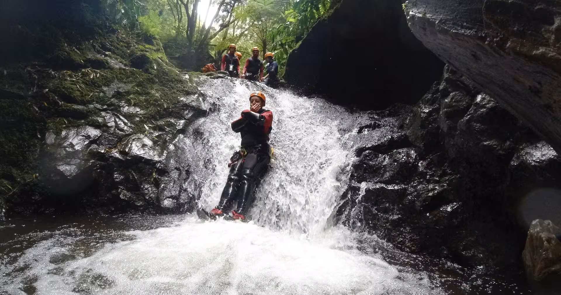 Canyoning adventurer sliding down a rocky waterfall chute into a pool, with helmeted group on guided tour