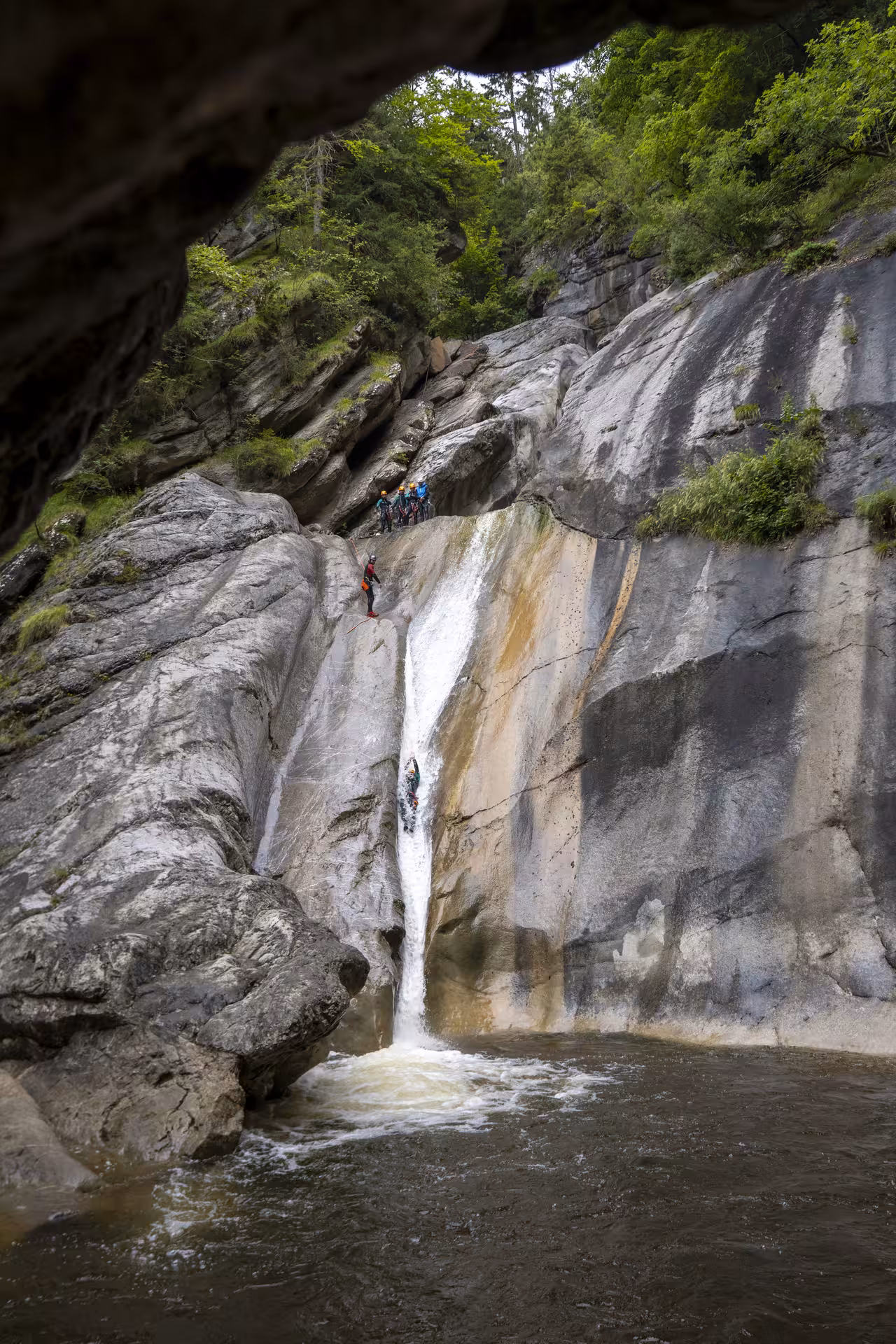 Adventurers canyoning down a steep waterfall at Chli Schliere near Luzern, surrounded by lush greenery and rugged rocks.