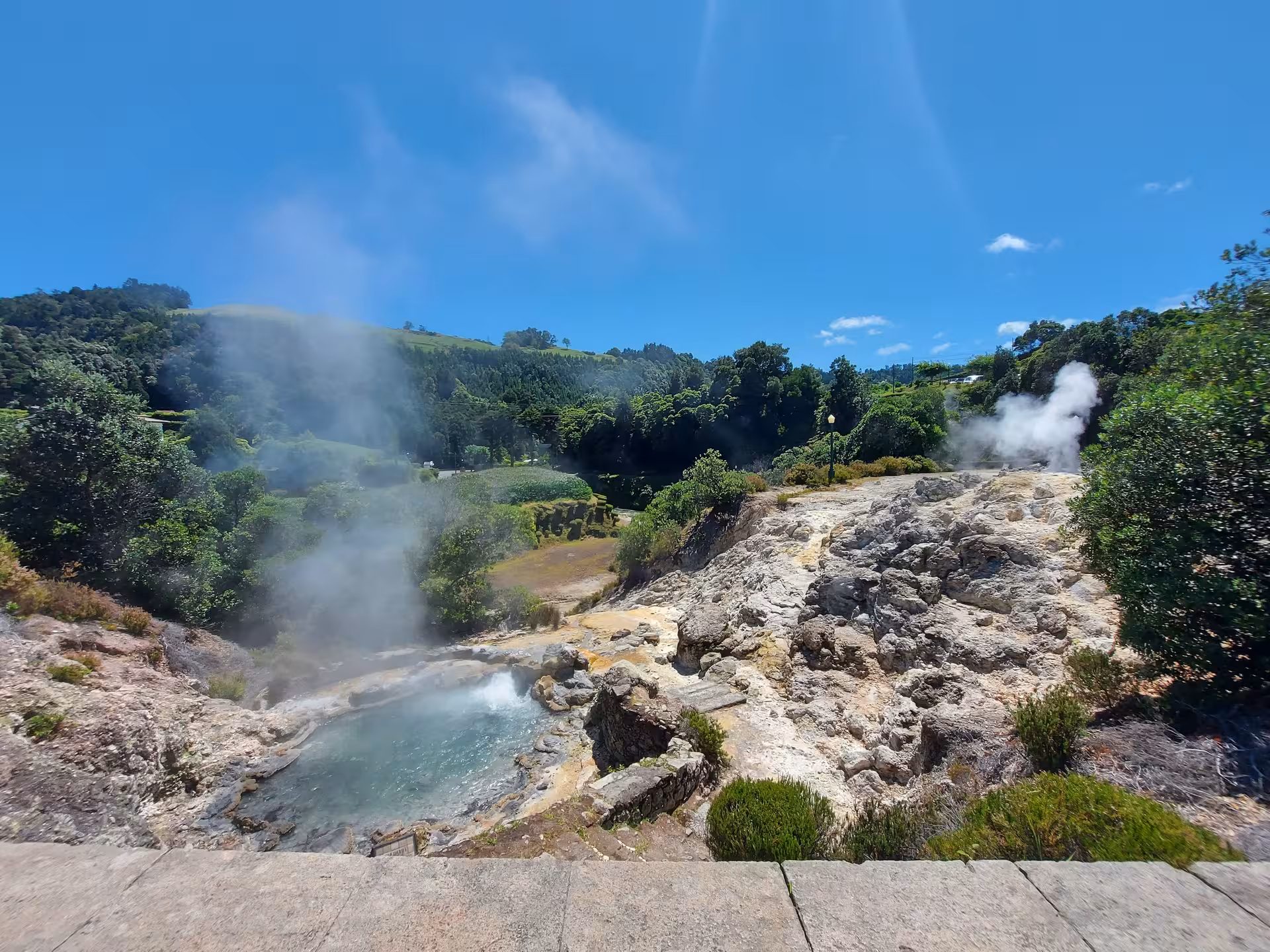 Steaming geothermal pool and rocky fumaroles in sunny Furnas, Azores, surrounded by green hills and forest