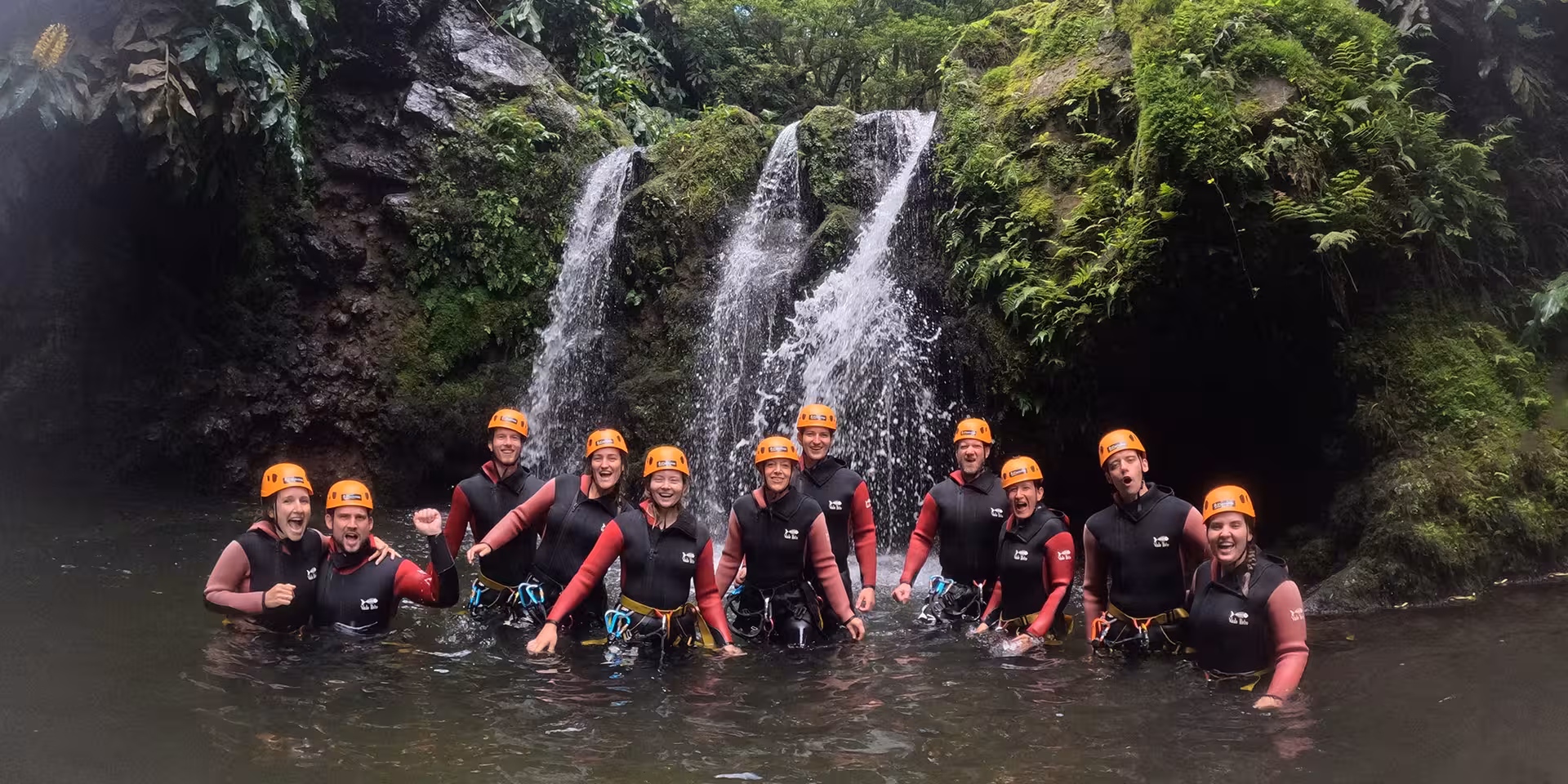 Smiling canyoning group in helmets and wetsuits standing in a natural pool under twin waterfalls on a guided tour