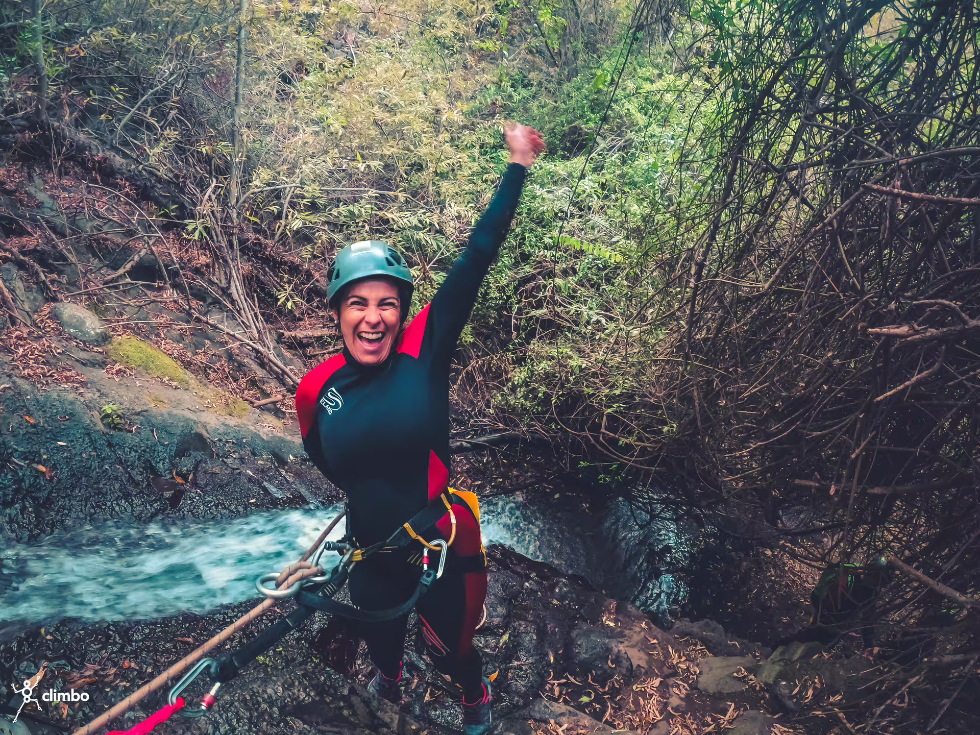 Excited canyoner raises fist triumphantly while navigating a waterfall in a vibrant rainforest setting.