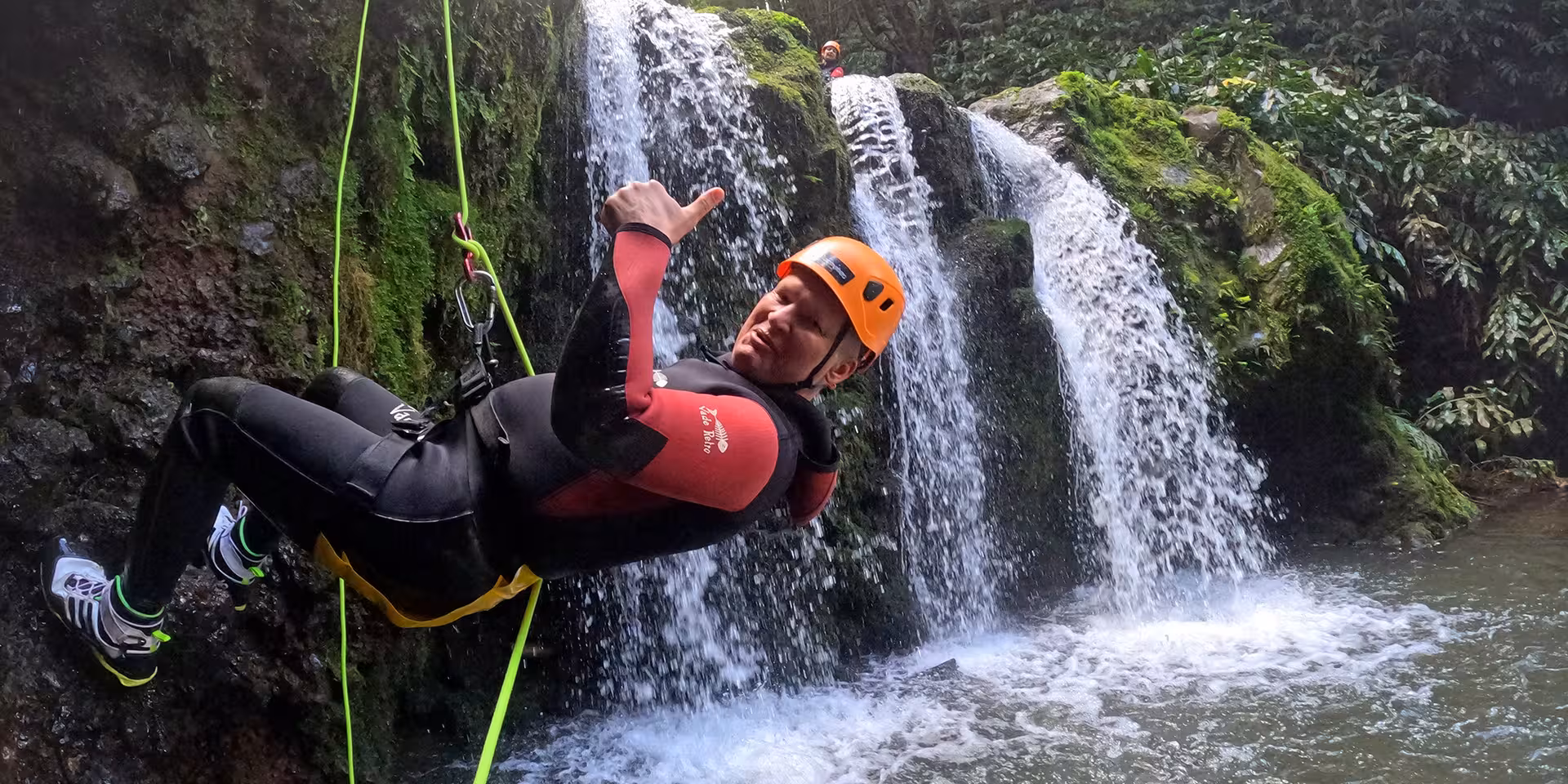 Canyoning rappel beside a mossy waterfall, helmeted adventurer on rope in wetsuit, guided canyon tour