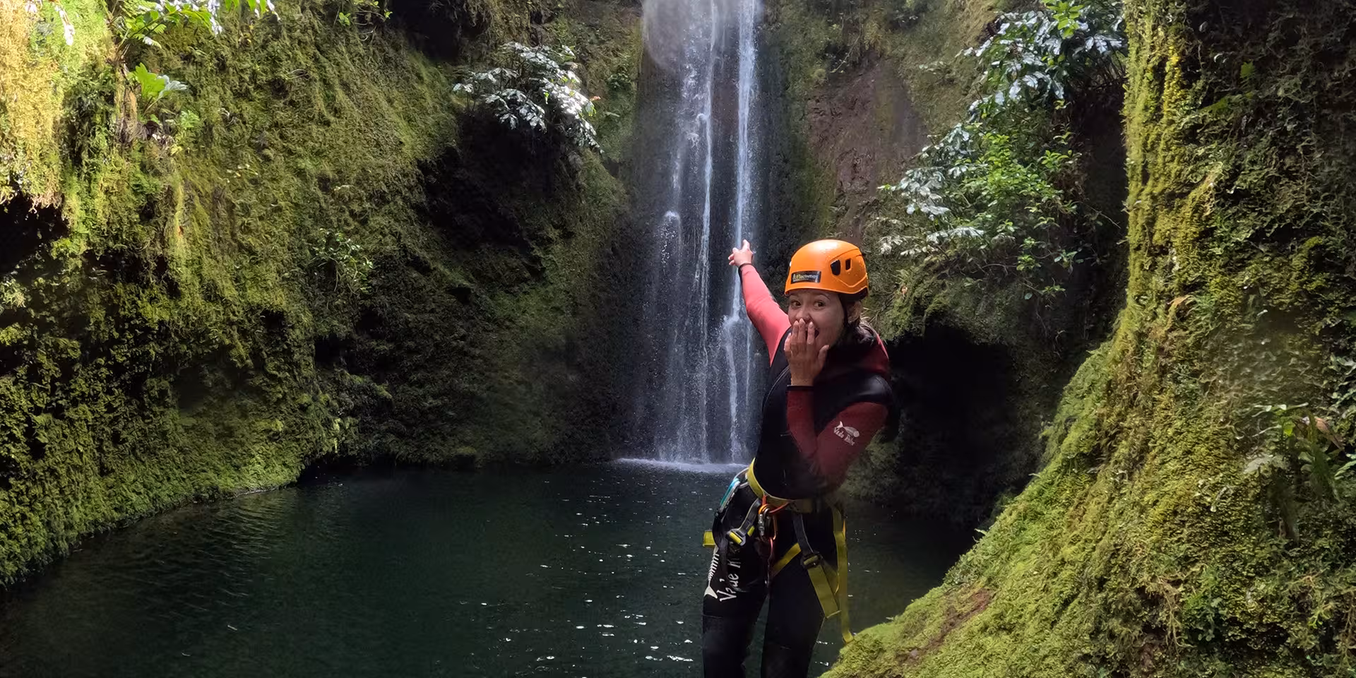 Canyoning Outward Shore guide in helmet by lush canyon waterfall pool, adventure rappel and swim tour