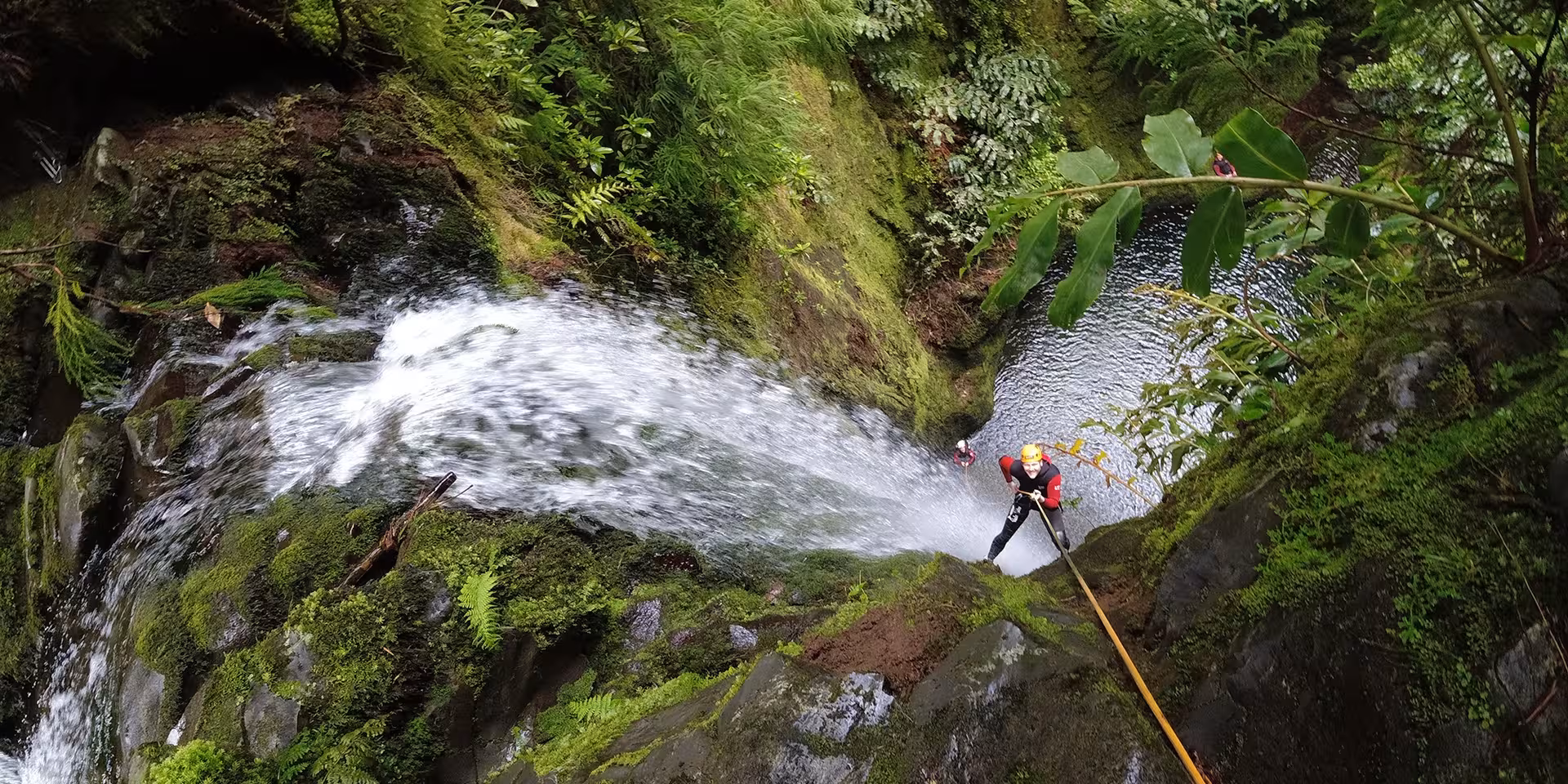 Canyoning Outward Shore rappel beside a rushing waterfall into a mossy gorge pool on a guided adventure tour