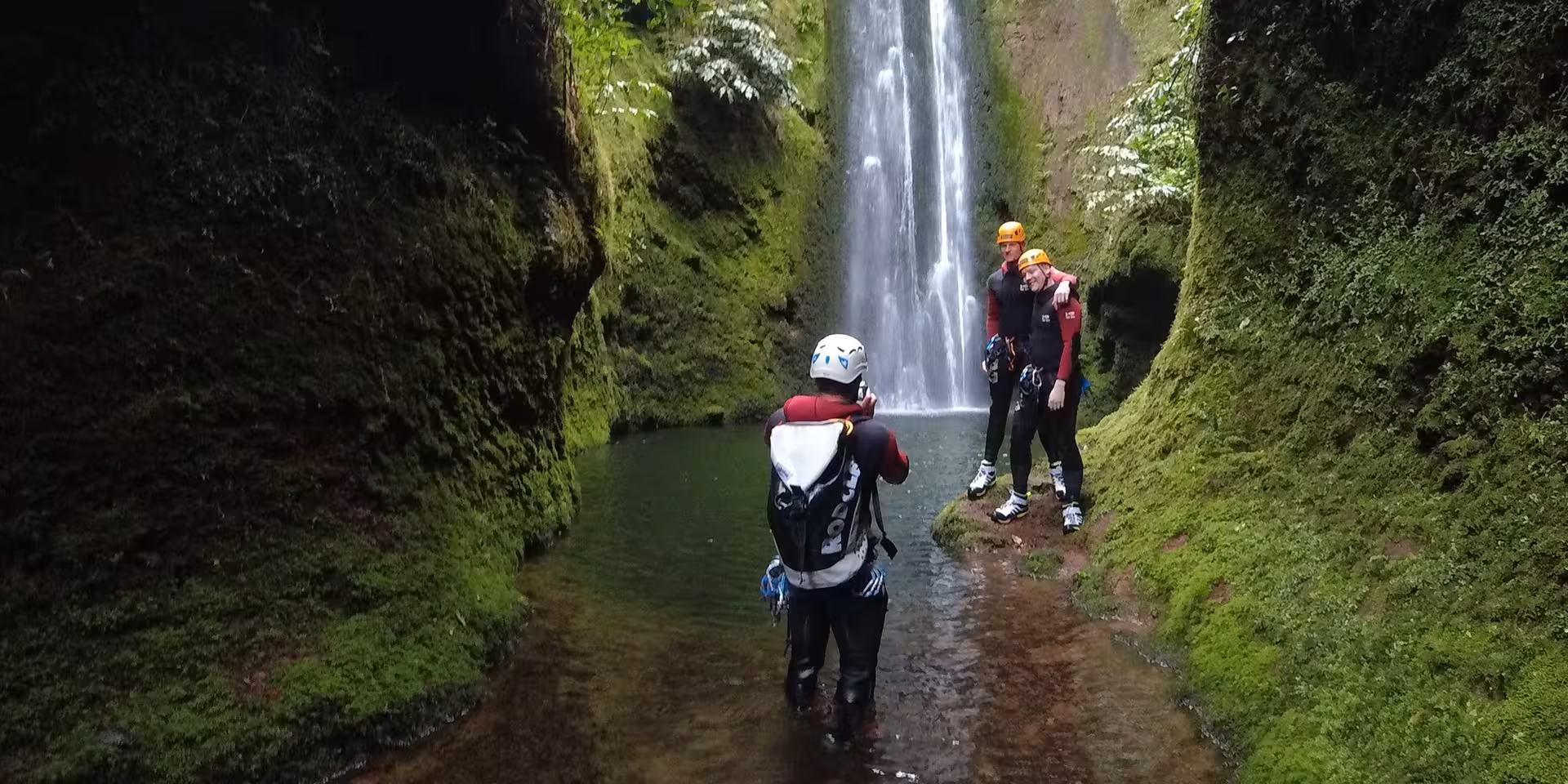 Canyoning group in wetsuits by tall waterfall in mossy gorge on CANYONING Outward Shore adventure tour