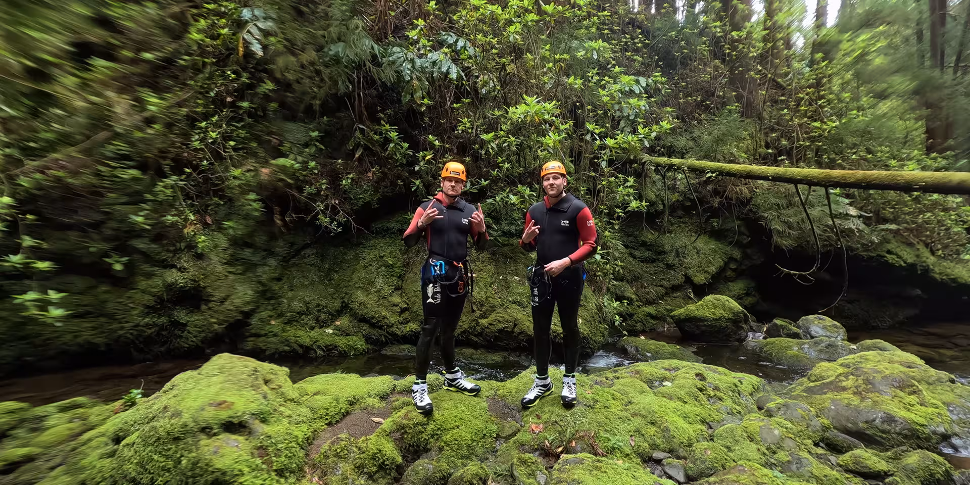 Canyoning Outward Shore participants in wetsuits and helmets on mossy river rocks, rainforest canyon trek