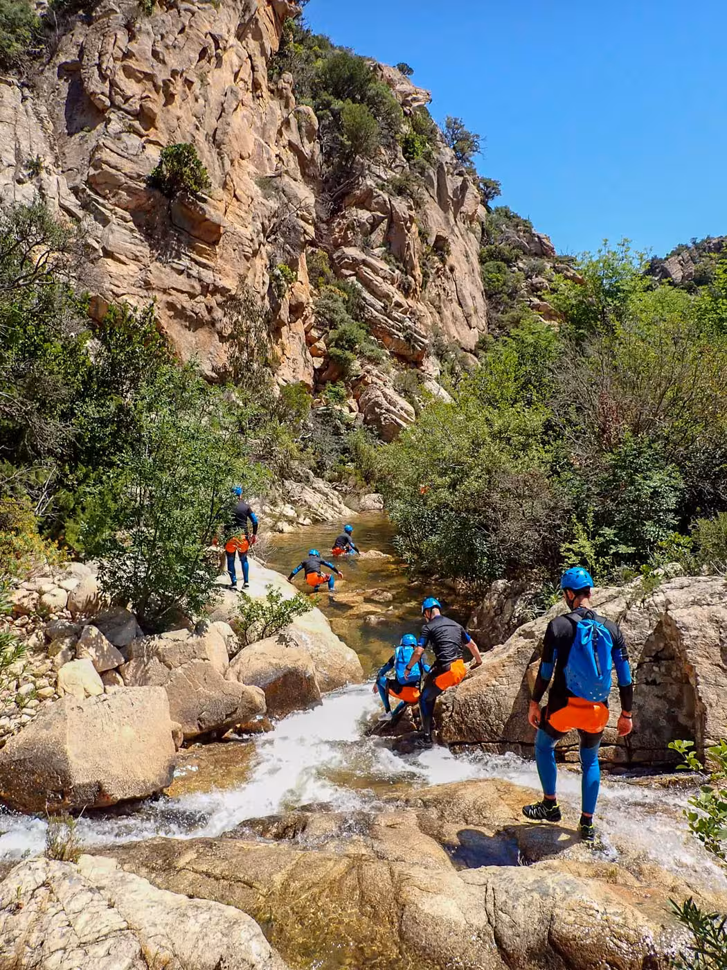 Group of canyoners navigating rocky stream in Rio Pitrisconi, San Teodoro, amidst striking cliffs and vibrant foliage.