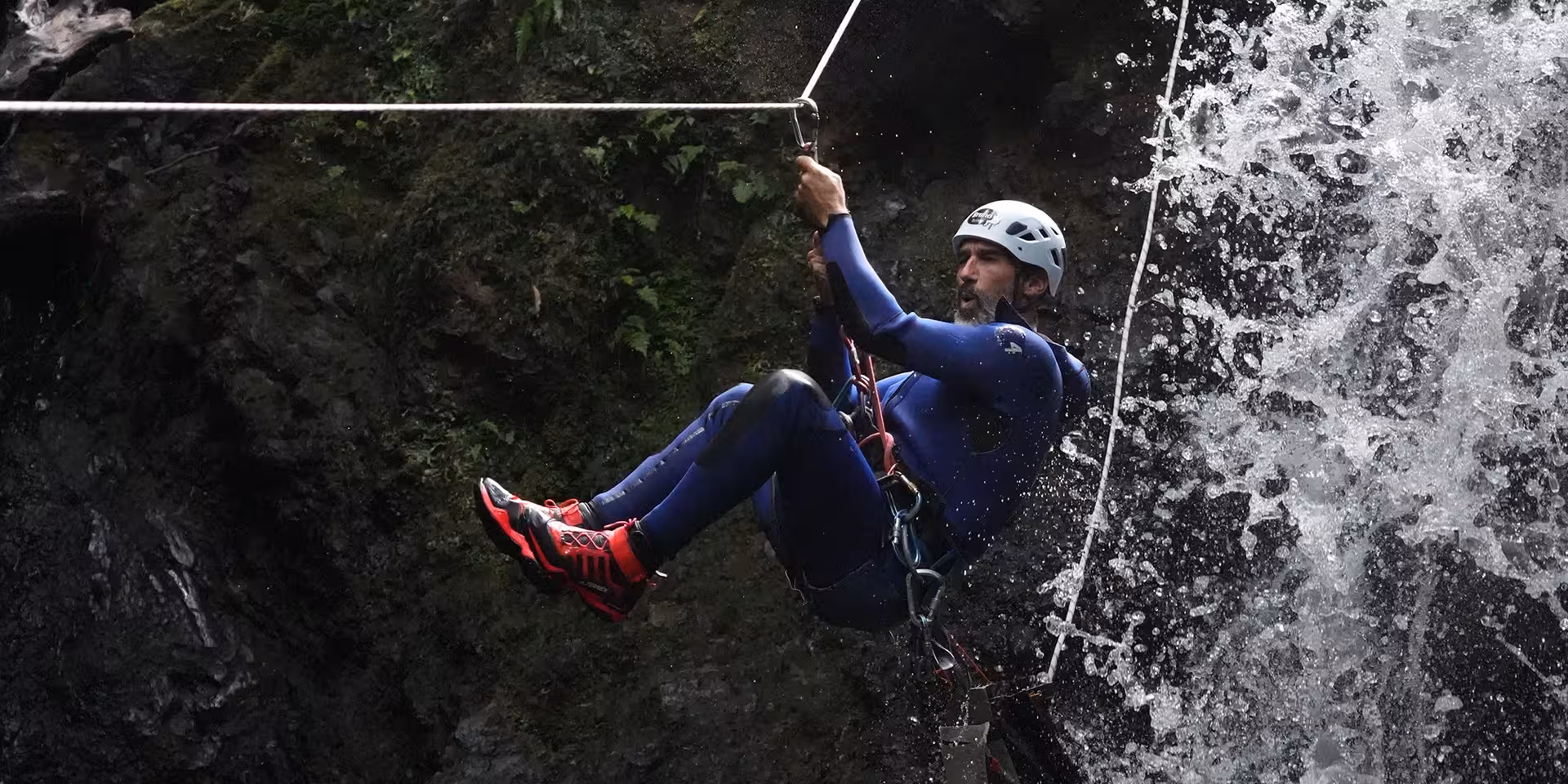 Canyoning zipline traverse through waterfall spray, climber in harness and helmet on rope, adrenaline adventure