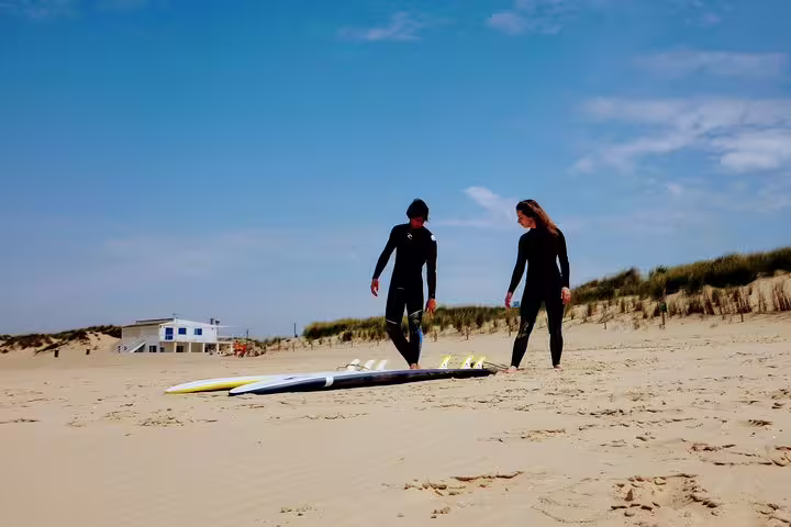Surfers in wetsuits prepare on Caparica Beach, Portugal, with surfboards on sand under a clear blue sky for a surf experience.