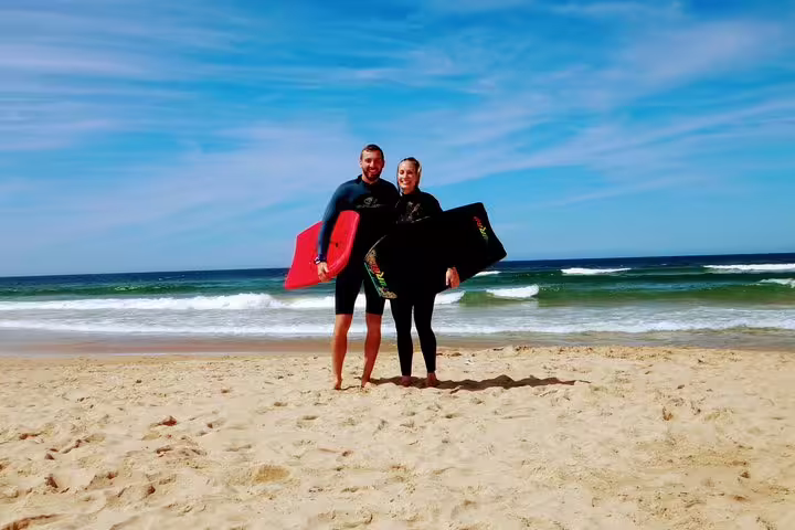Couple enjoying a Caparica bodyboarding adventure on a sunny beach with clear blue skies and ocean waves in the background.