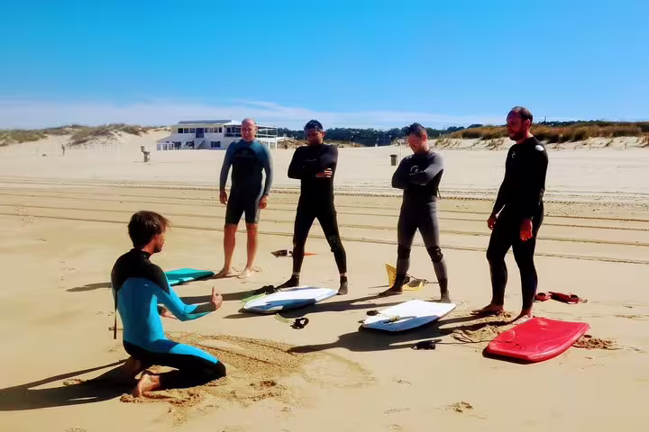 Surf instructor teaching a group on Caparica Beach, Portugal, with surfboards in the sand under clear blue skies.