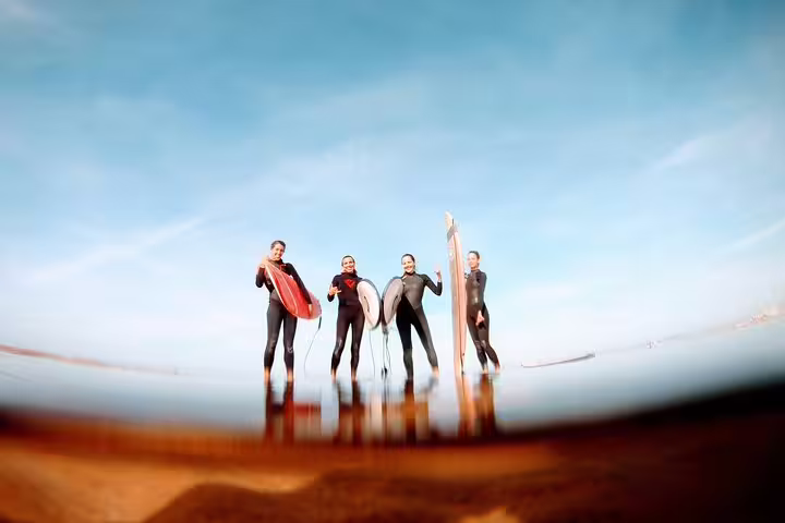 Group of surfers in wetsuits holding boards on Caparica beach under a clear sky, ideal for surf and yoga enthusiasts.