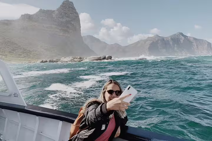 Tourist capturing a selfie on a boat with stunning Cape Peninsula mountains and ocean views in the background.