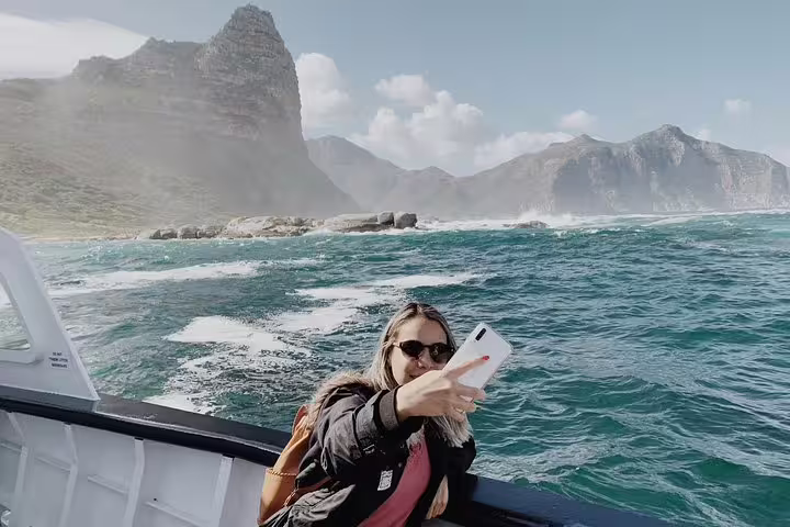 A tourist takes a selfie on a boat with scenic views of the ocean and rugged cliffs near the Cape of Good Hope.