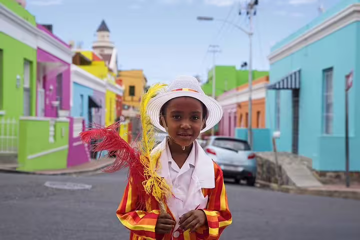 A child in colorful attire and a hat holds feathers, standing in the vibrant Bo-Kaap neighborhood in Cape Town.
