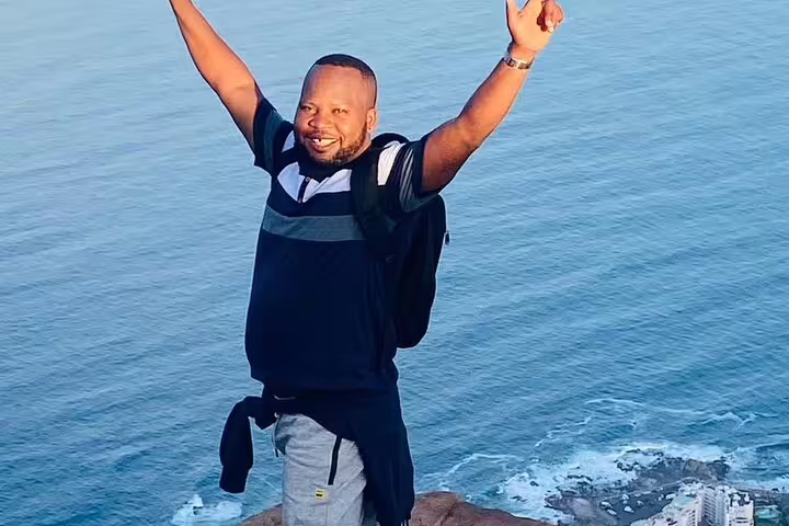 Excited man with arms raised stands atop a cliff with ocean view, celebrating Cape Town paragliding adventure.