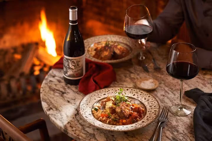A cozy dining setup with red wine and gourmet dishes by a fireplace, representing a wine tour in Cape Town's Winelands.