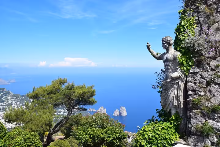 Statue overlooking Capri's stunning landscape and Faraglioni rocks, surrounded by lush greenery and blue skies.