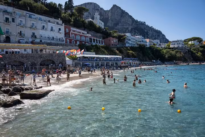 Crowded beach in Capri with tourists enjoying the sun, near vibrant seaside buildings and clear azure waters.