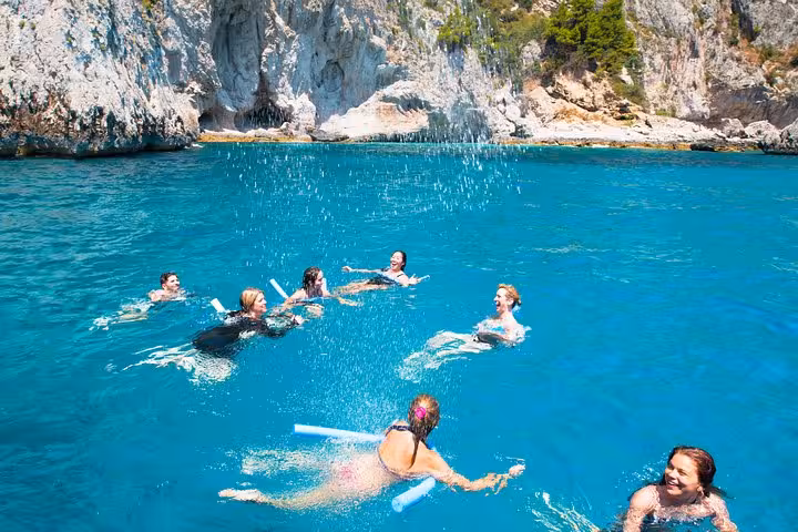 Group of tourists swimming with noodles in the turquoise waters near Capri's rocky coastline.