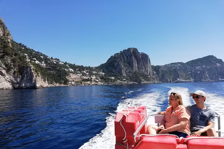 Couple enjoying a boat tour with scenic views of Capri's coastline and Faraglioni rocks under a clear blue sky.