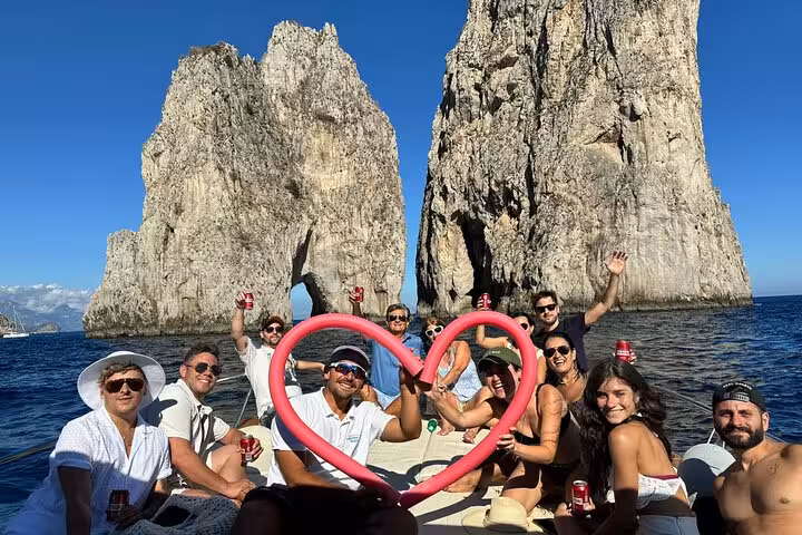 Group of friends celebrating with drinks and heart-shaped float in front of Capri's iconic sea stacks.