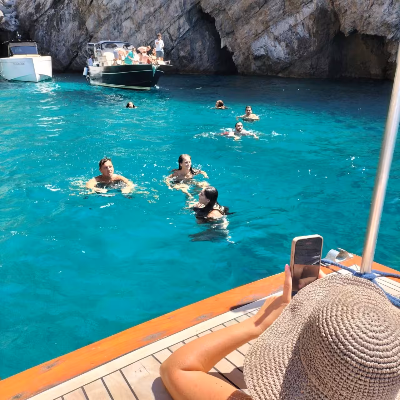 Swimmers in turquoise cove near Capri grotto as guests relax on boat deck, Capri one-day boat tour experience