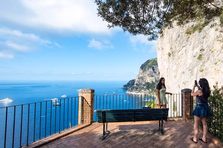Tourists capturing photos at a scenic viewpoint overlooking the stunning coastline of Capri with clear blue sea.