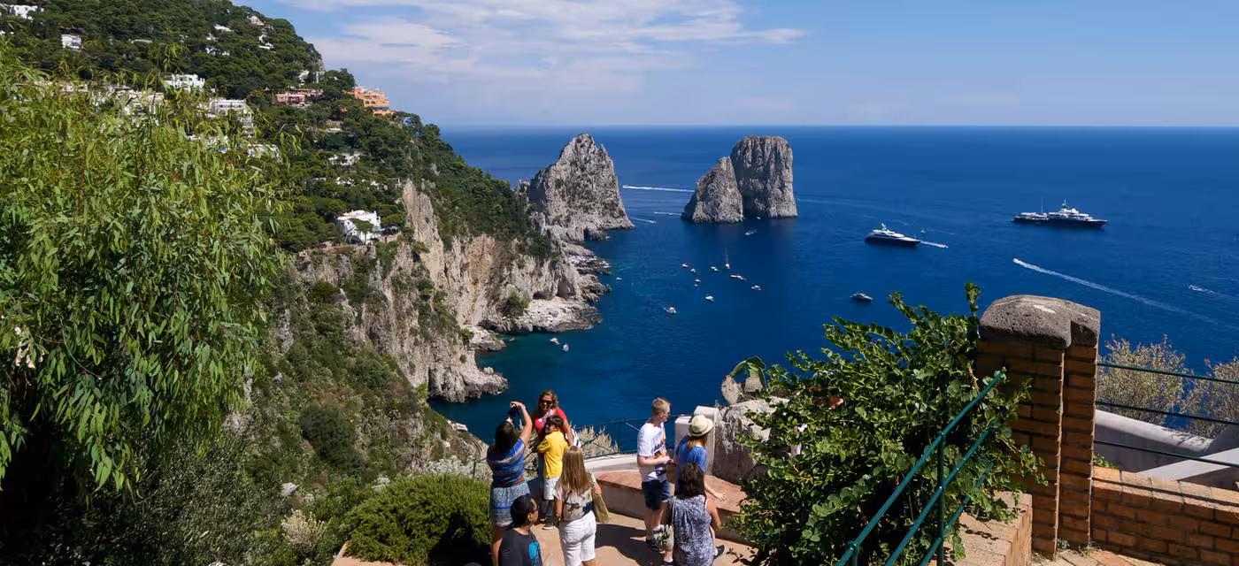 Tourists enjoy stunning views of Faraglioni rock formations from a scenic overlook in Capri, Italy.