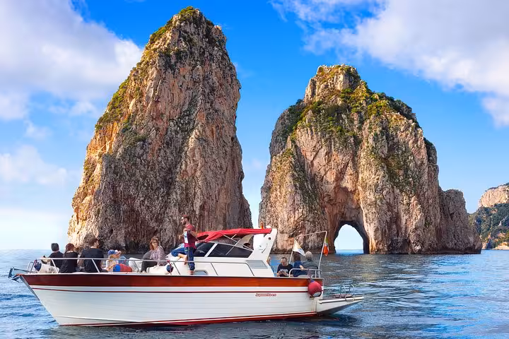 A boat full of tourists sails near the iconic Faraglioni rock formations during a Capri Island day trip.