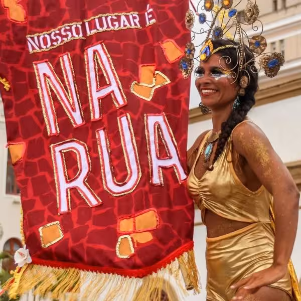 Carnival performer in gold costume with decorative headpiece, standing by a festive street party banner.