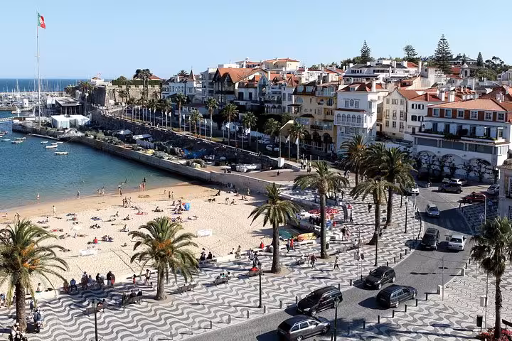 Aerial view of Cascais beach with sunbathers, palm-lined promenade, and picturesque seaside townscape.