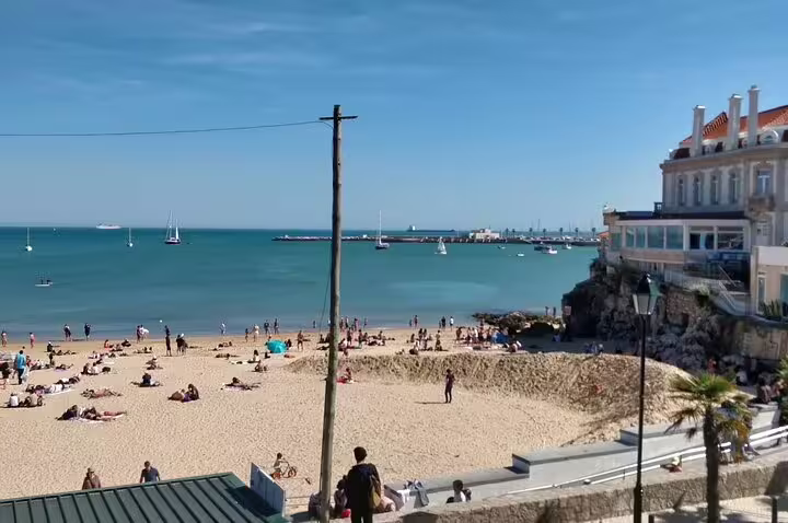 Scenic view of a sunny beach in Cascais with people relaxing, sailboats in the water, and a historic building by the shore.