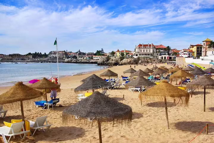 Sunny beach in Cascais with straw umbrellas and lounge chairs, part of the Sintra Cabo da Roca Cascais and Estoril tour.
