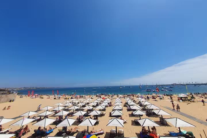 Sunny beach in Cascais with rows of umbrellas, blue ocean, and boats, part of the Sintra, Cabo da Roca & Cascais tour.