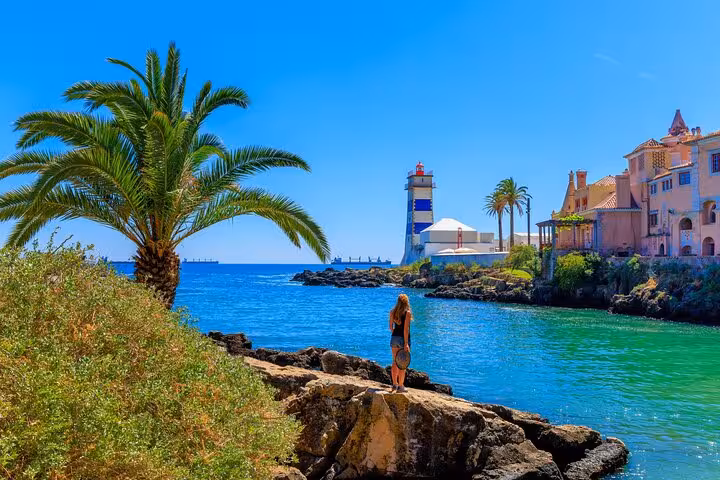 Vibrant view of Cascais with a person admiring the coastal lighthouse, palm trees, and turquoise waters under a sunny sky.