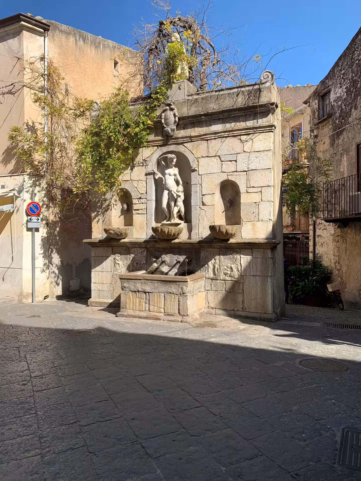 Historic stone fountain with classical statues and ivy in a narrow medieval street of Castelbuono on a private tour from Palermo