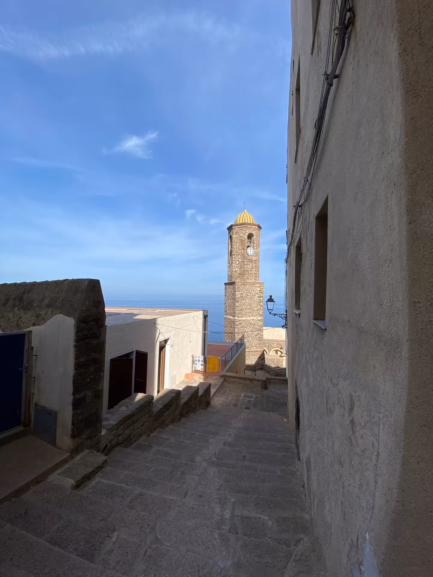 Charming medieval alley in Castelsardo with view of historic clock tower under a clear blue sky.