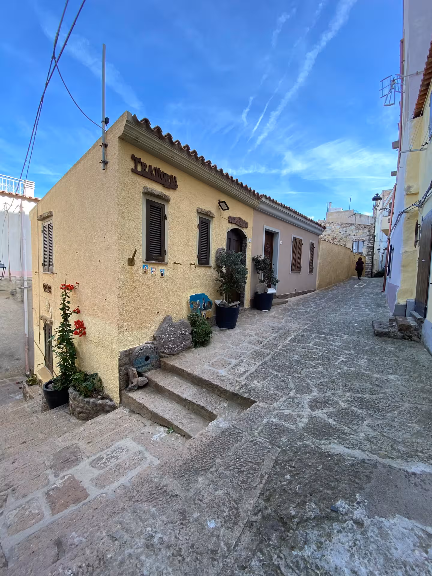 Charming narrow street in Castelsardo with traditional buildings and rustic trattoria under a clear blue sky.