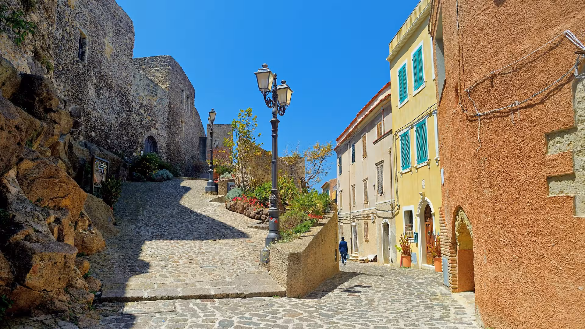 Charming cobblestone street in Castelsardo old town with historic buildings and vibrant flowers under a clear blue sky.