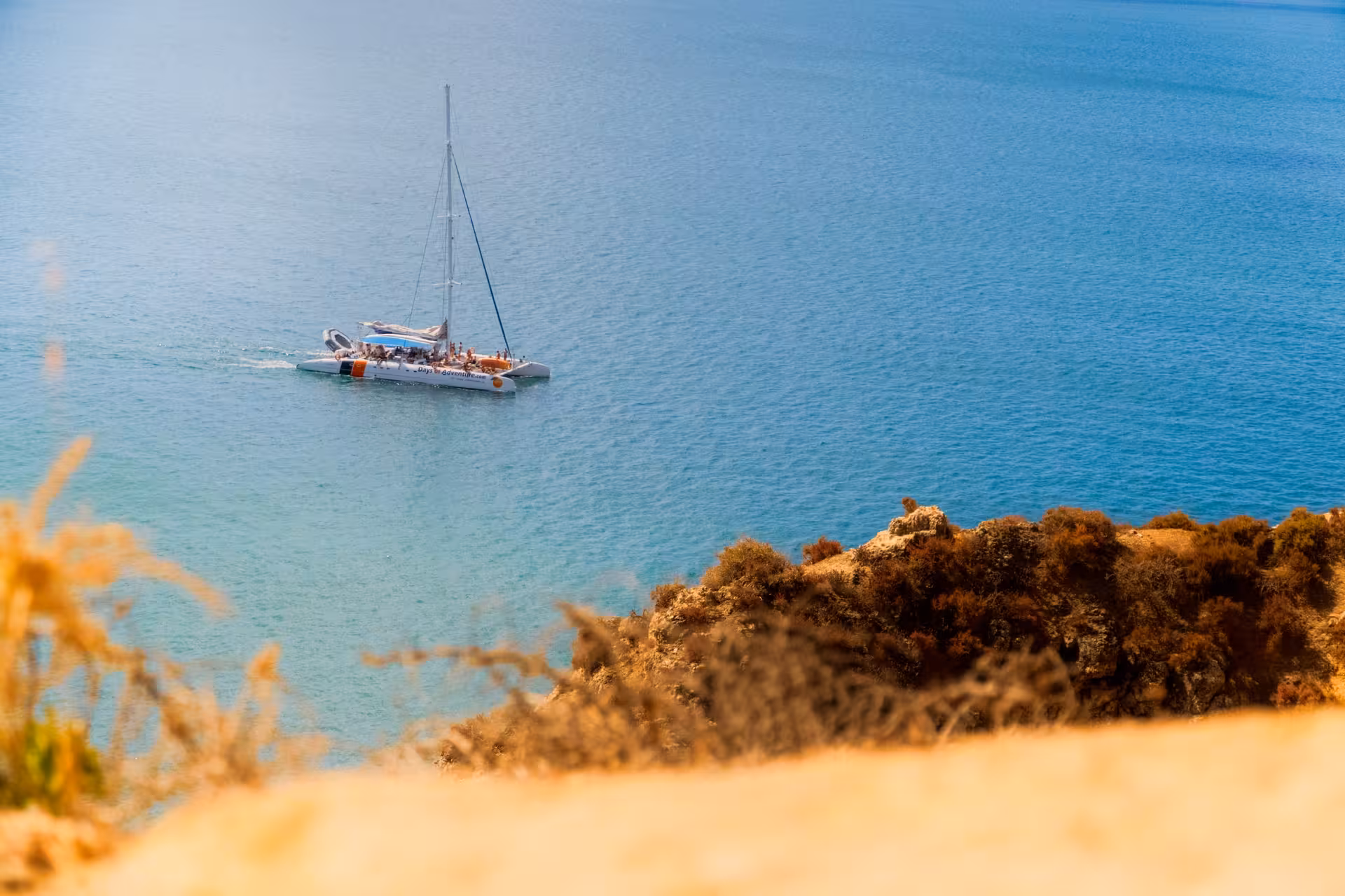 Aerial cliffside view of a catamaran sailing the turquoise Lagos Algarve waters on a sunny boat tour with music.