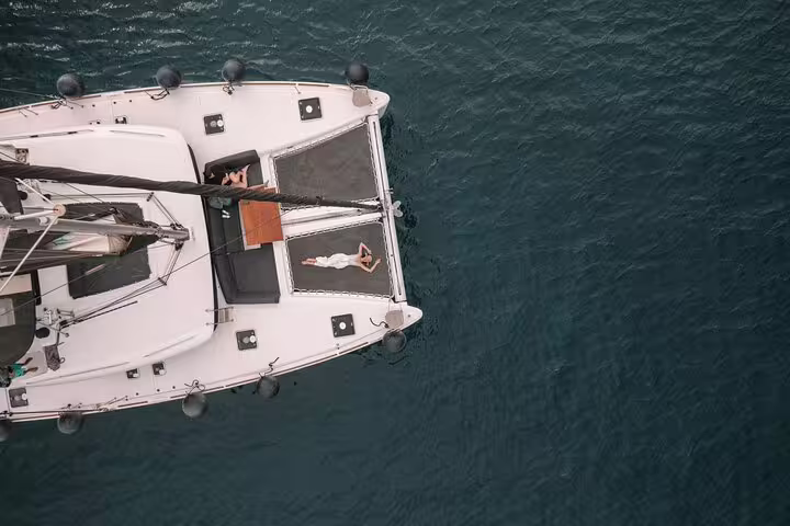 Aerial view of a catamaran with a guest relaxing on deck, enjoying a private cruise in Santorini's tranquil waters.