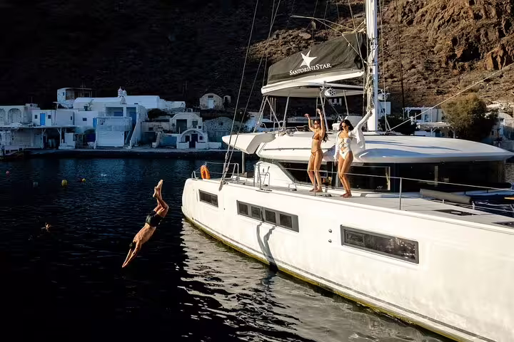 Guests diving from a luxurious catamaran during a Santorini cruise with scenic views and onboard excitement.