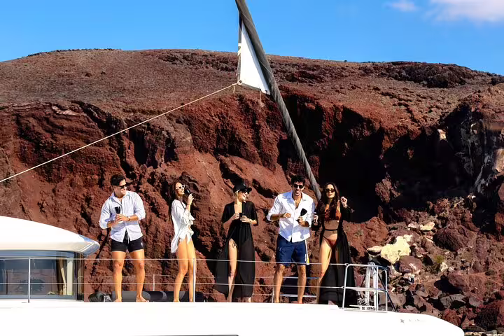 Group enjoying drinks on a catamaran with stunning views of Santorini's red cliffs during a half-day cruise.